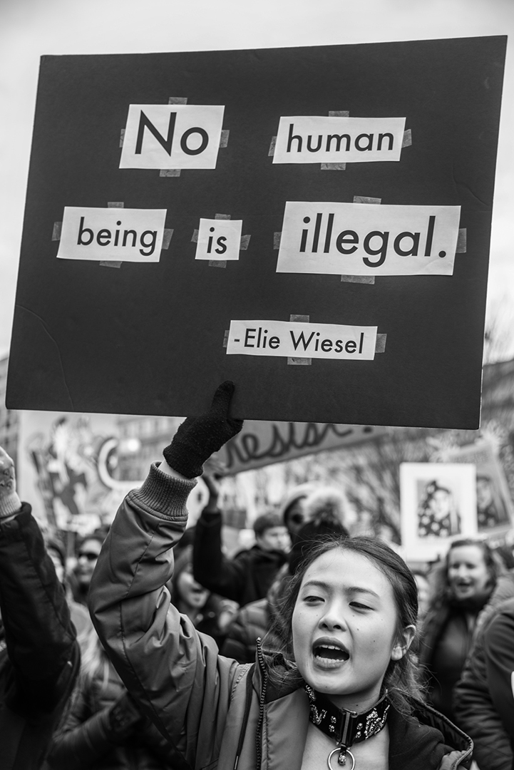 A young woman of Asian decent standing in a crowd of protesters holds a sign above her head that reads "No Human Being Is Illegal - Elie Wiesel." This documentary photography was created by Kaitlin K Walsh Photography.